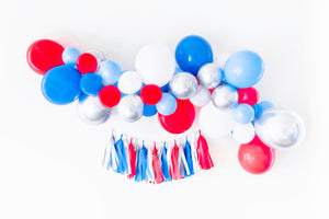 A perfectly patriotic themed balloon garland displayed on a white wall with a themed tassel garland hanging underneath. Balloons are a mix of red, blue, light blue, chrome silver, and white. Tassels are in matching red, white, blue, and silver.