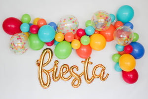 A gold mylar fiesta script balloon is hung on the wall underneath a colorful latex balloon garland.