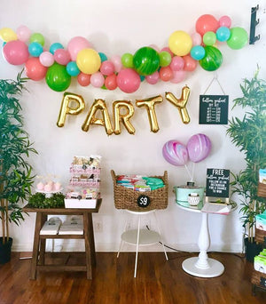 Gold balloon letters hang from a wall and spell PARTY. There is a colorful garland hanging above it.
