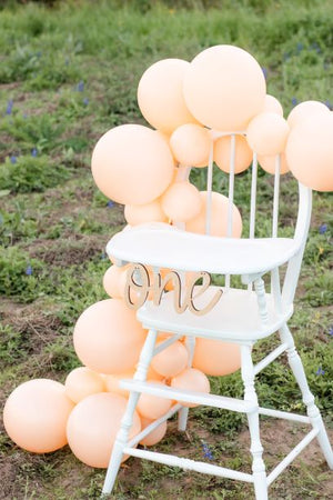 A white high chair sits in a field with a wooden cursive banner that says one hanging in front. There is a blush/peach colored balloon garland draped over the back and down the side of the highchair all the way to the ground.