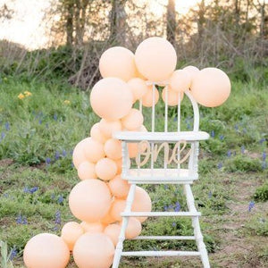 A white high chair sits in a field with a wooden cursive banner that says one hanging in front. There is a blush/peach colored balloon garland draped over the back and down the side of the highchair all the way to the ground.