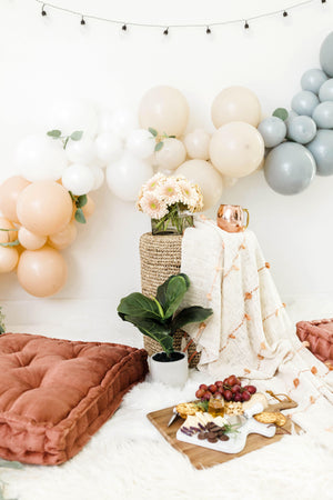 A close up shot shows a full charcuterie board on the ground in front of the garland as well as a few soft flowers and white rug.