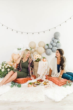 Two women sit on the floor in front of the balloon garland looking at each other and laughing.