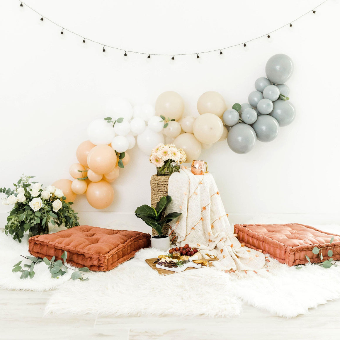 A beautiful boho themed garland drapes across a white wall. The balloons are gathered together by color in order from left to right. Colors are blush, white, tan, and grey.