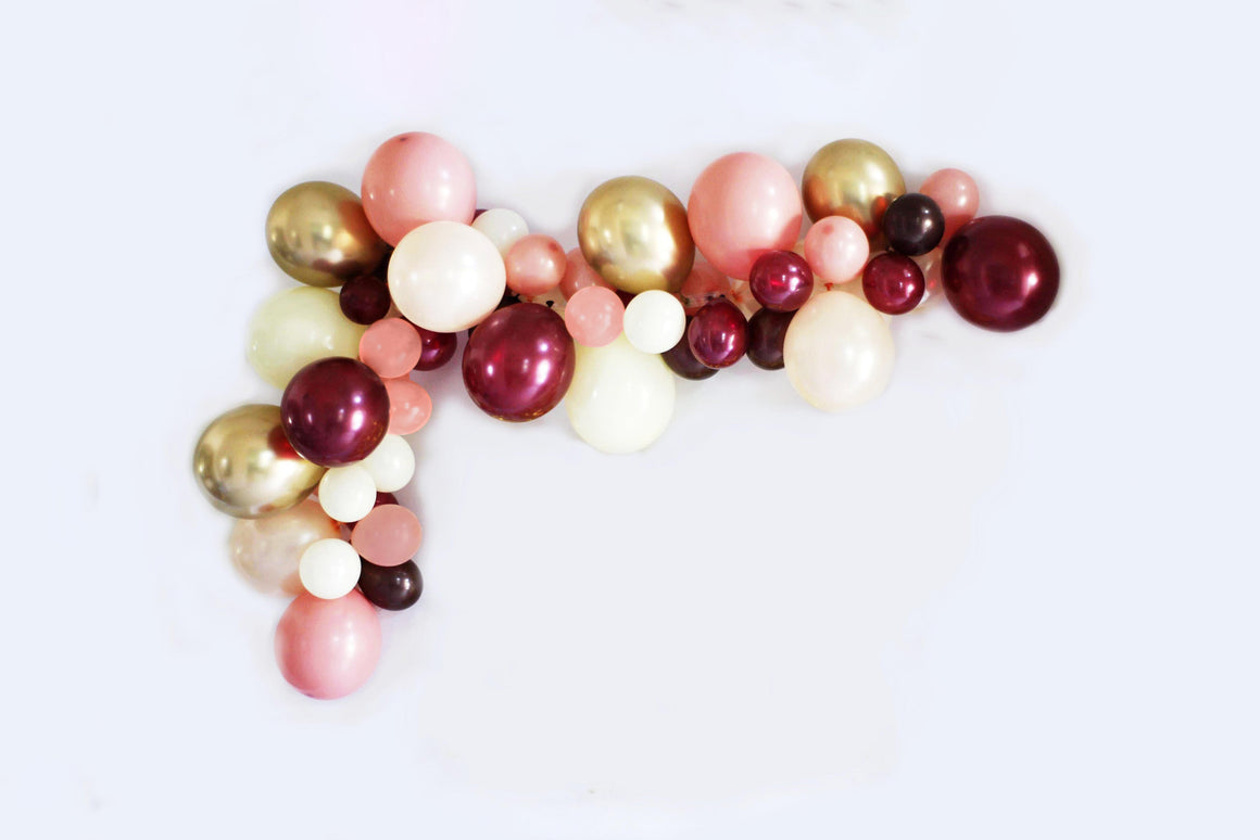 Balloon garland with the colors pearl burgundy, pearl peach, ivory, chrome gold, mauve, and brown hangs from a white wall.