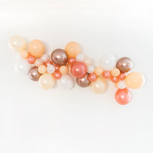 A front view of a bridal themed balloon garland is draped across a white wall. The balloon garland consists of various 5 inch and 11 inch sized balloons in the colors of pearl white, rose gold, blush, pearl peach, and chrome rose gold.