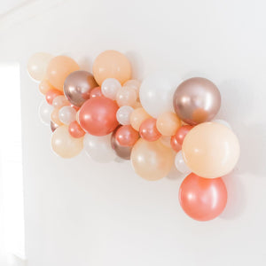 A side view of a bridal themed balloon garland is draped across a white wall. The balloon garland consists of various 5 inch and 11 inch sized balloons in the colors of pearl white, rose gold, blush, pearl peach, and chrome rose gold.
