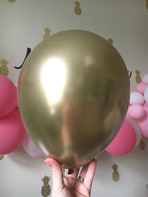 A close up view shows the gold chrome latex balloon inflated with a pretty pink balloon garland in the background.