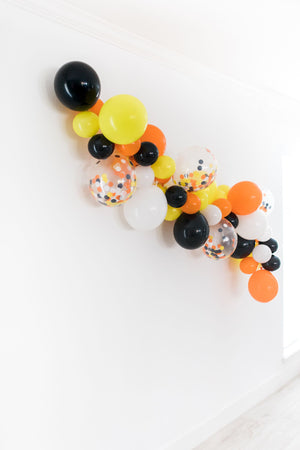 A side view of the balloon garland on a white wall to show another angle. With the colors shown of orange, white, black, and yellow. As well as a few 11 inch clear confetti filled matching balloons.