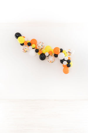 Another straight on view of the balloon garland on a white wall. With the colors shown of orange, white, black, and yellow. As well as a few 11 inch clear confetti filled matching balloons.