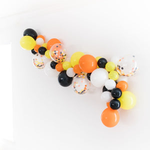 A side view of a construction balloon garland strung on a white wall. The balloon garland displays the various 5 inch and 11 inch balloons in the colors of orange, yellow, black, and white. As well as a few matching confetti filled clear balloons.