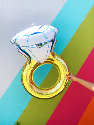 A womans hand is holding a jumbo diamond ring balloon in front of a colorful wall.