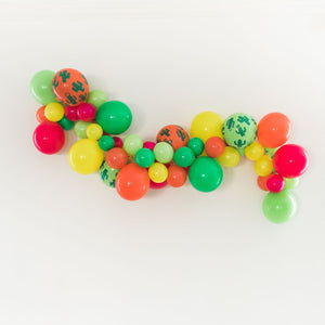 A fiesta themed balloon garland is hanging from a white wall. Colors shown are yellow, spring green, lime, coral, and wildberry in both 5 inch and 11 inch sizes. As well as a few 11 inch cactus print balloons sprinkled in.