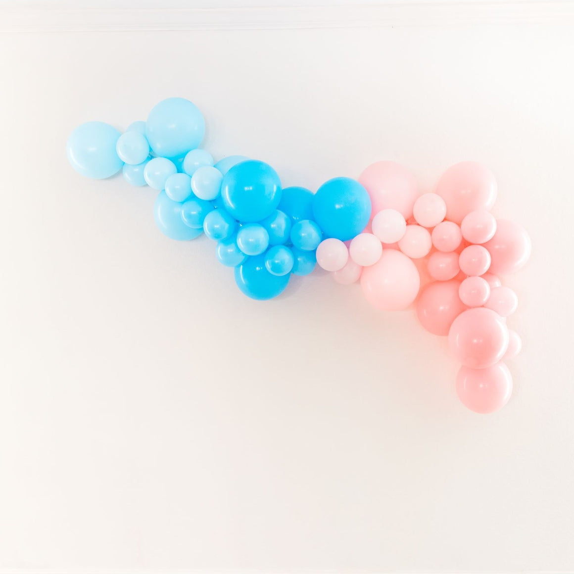 A soft pretty balloon garland is draped on a white wall. With a mix of pastel light blue, light blue, pastel pink, and pink balloons in that order from left to right.