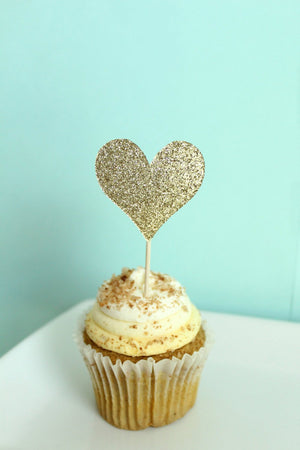 A cupcake with a gold glitter heart topper sits on top of a white table.