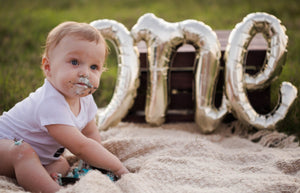 Little boy with cake on his hands and face sitting in front of a cursive white gold mylar ONE balloon.