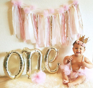 Little girl wearing a princess crown sitting next to a 30 inch by 16 inch white gold cursive ONE balloon.