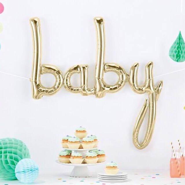Gold script baby balloon hanging from a string against a white wall.