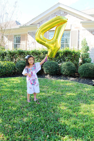 A little girl wearing a dress standing in a yard holding a helium filled number four gold colored balloon.