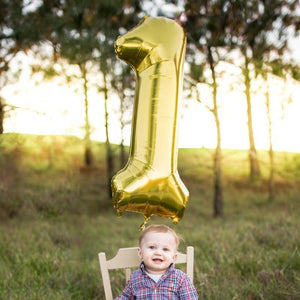 A one year old boy sitting in a field with a gold number one balloon.