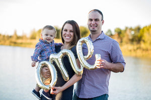 Couple holding a toddler with a cursive white gold ONE mylar balloon.