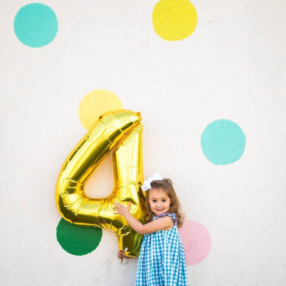 A little girl holding a gold 4 balloon standing in front of a polka dot background.