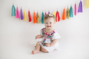 A baby girl sits smiling in front of a hot pink, purple, yellow, red, orange, gold, and teal tissue tassel garland strung across the wall behind her.