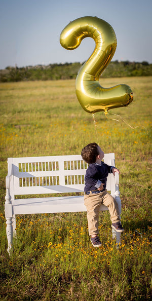 A two year old boy sitting on a white bench in a field looking up at a gold helium filled number two balloon floating.