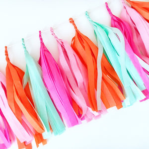 A arial shot of a hot pink, pink, orange, and mint tissue tassel garland is taken of tassels strung on twine and laying on a white table. The tassels are approximately nine inches long.