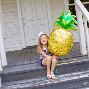A little girl sits on a porch holding a giant 37 inch pineapple balloon.