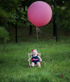 A toddler baby girl is sitting in a pink rocker in a grass field with a jumbo 36 inch pink balloon tied on the rocker.