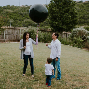 Couple standing in their backyard with a toddler and holding a black jumbo balloon filled with either pink or blue confetti.