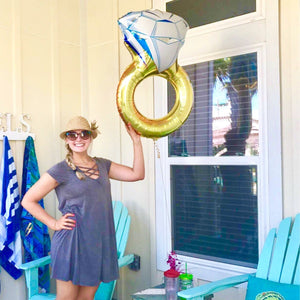 A woman smiling is outside holding a diamond ring balloon.