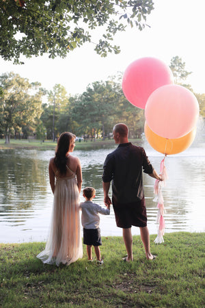 Couple holding their little sons hand are standing in front of a lake. The man is holding three 36 inch balloons.