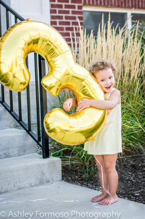 A little kid standing in the grass holding up a gold number three balloon.