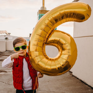 A little boy throwing the peace sign holding a jumbo 6 balloon.