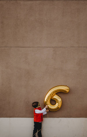 A little boy in front of a cement wall wearing a red vest and holding a giant 6 gold balloon.