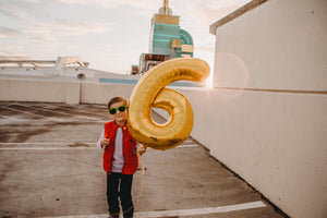 A little boy smiling outside holding a gold big 6 balloon.