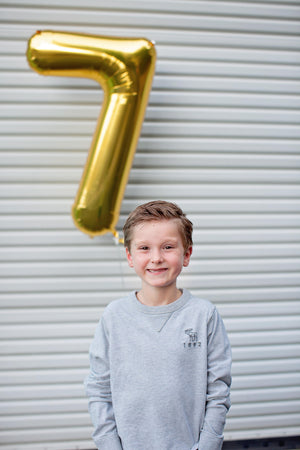 A boy smiling in front of a white colored garage door with a helium filled gold number seven balloon floating behind him.