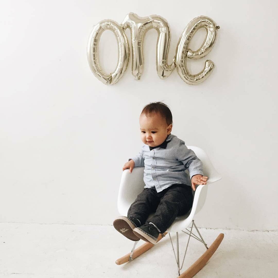 Little boy sitting in white rocker with cursive white gold ONE mylar balloon hanging on the wall.