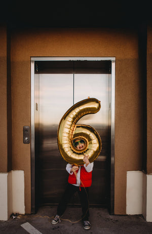 A little boy standing in front of an elevator holding a jumbo gold mylar number six balloon.
