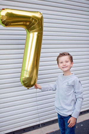 A boy smiling in front of a white colored garage door while holding a gold number seven balloon at his side.