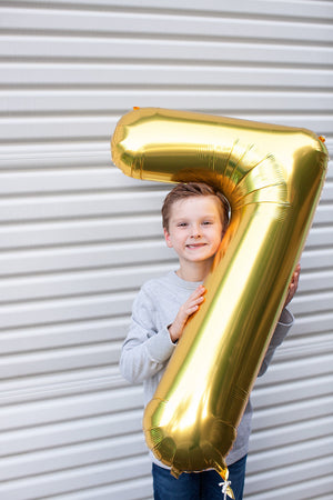 A boy smiling in front of a white colored garage door holding a gold number seven balloon that is almost as big as he is.