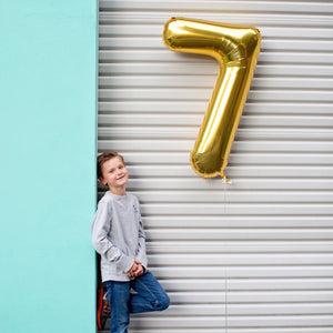 A boy smiling and leaning against a wall outside posing next to a floating gold number seven balloon.