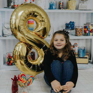 A smiling girl sitting next to an inflated gold number eight balloon in front of a bunch of colorful filled candy jars.