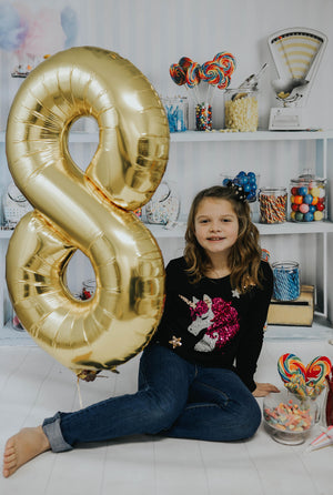 A girl sitting next to an inflated gold number eight balloon with a bunch of colorful candy filled jars on shelves behind her.