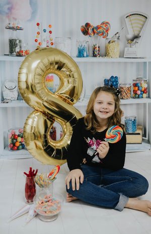A girl smiling sitting next to a gold number eight balloon while holding a jumbo colorful lollipop in her left hand.