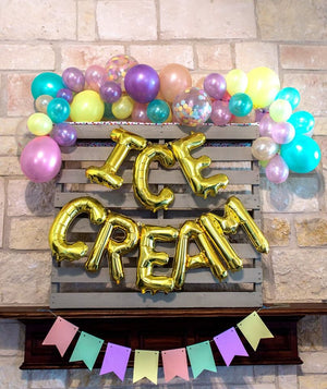 Gold mylar letter balloons spelling ICE CREAM surrounded by a balloon garland with the colors pink, purple, yellow, and blue and a matching banner.