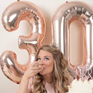 A woman sitting on the ground holding a cake and rose gold 30 balloons on the wall behind her.
