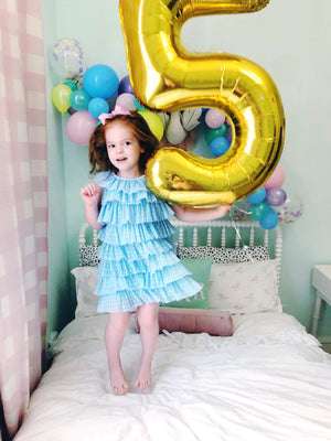 A little girl smiling and bounding on a white bed holding a gold number five balloon.